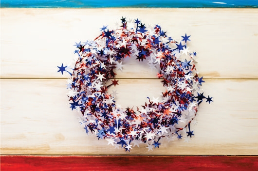 A wreath made of red, white, and blue star-shaped decorations hangs on a wooden background painted in matching colors.