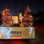 May show: A float decorated with lit Christmas trees and a "Rotary Club of Chatham" banner moves along a street at night.