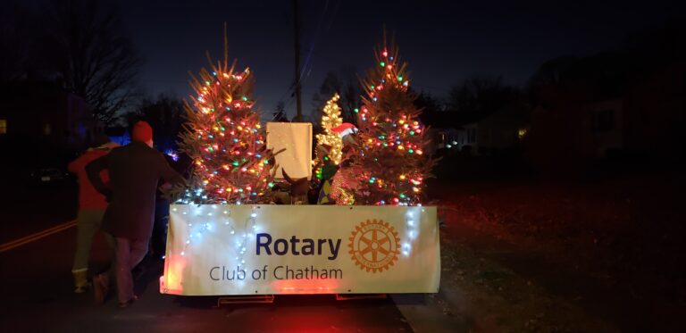 May show: A float decorated with lit Christmas trees and a "Rotary Club of Chatham" banner moves along a street at night.