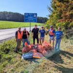 May show: Six people stand on grass by a rural road with orange trash bags and litter, under a blue "Adopt-A-Highway" sign for the Chatham Rotary Club.