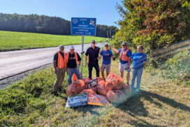 May show: Six people stand on grass by a rural road with orange trash bags and litter, under a blue "Adopt-A-Highway" sign for the Chatham Rotary Club.