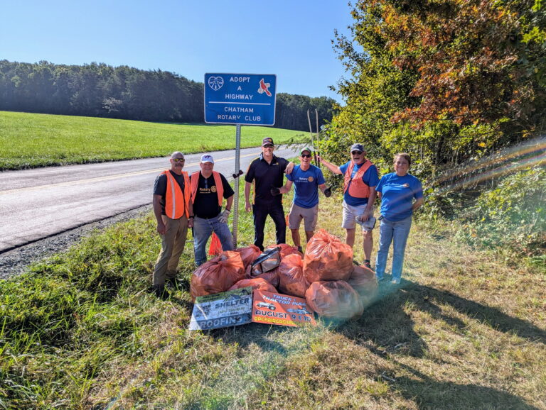 May show: Six people stand on grass by a rural road with orange trash bags and litter, under a blue "Adopt-A-Highway" sign for the Chatham Rotary Club.