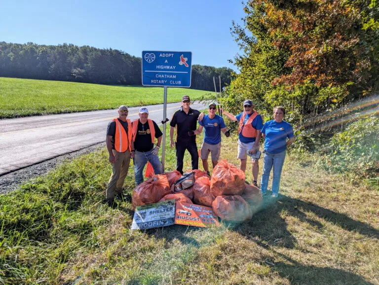 May show: Six people stand on grass by a rural road with orange trash bags and litter, under a blue "Adopt-A-Highway" sign for the Chatham Rotary Club.