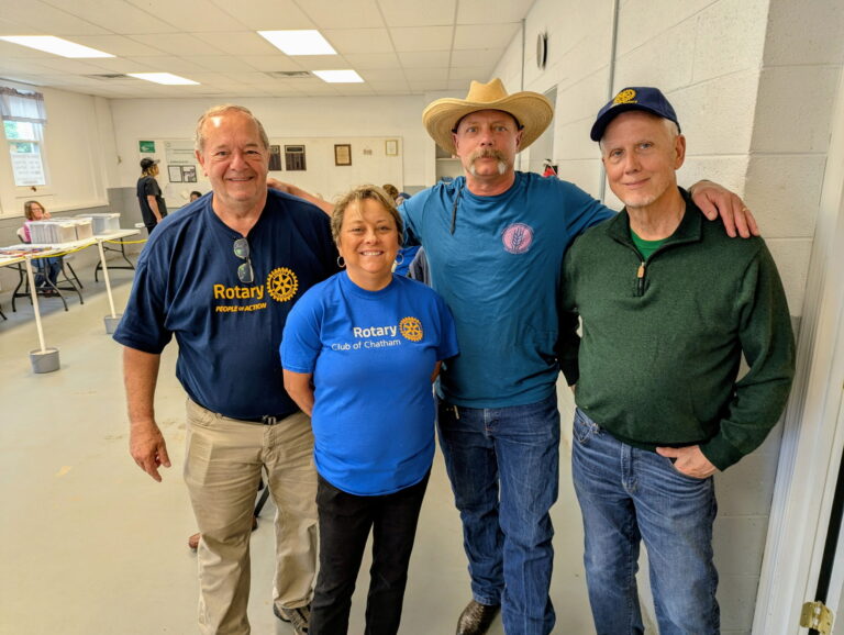 May show: Four adults stand together indoors, two wearing Rotary Club shirts, one in a cowboy hat, and one in a green sweater, posing for a group photo.