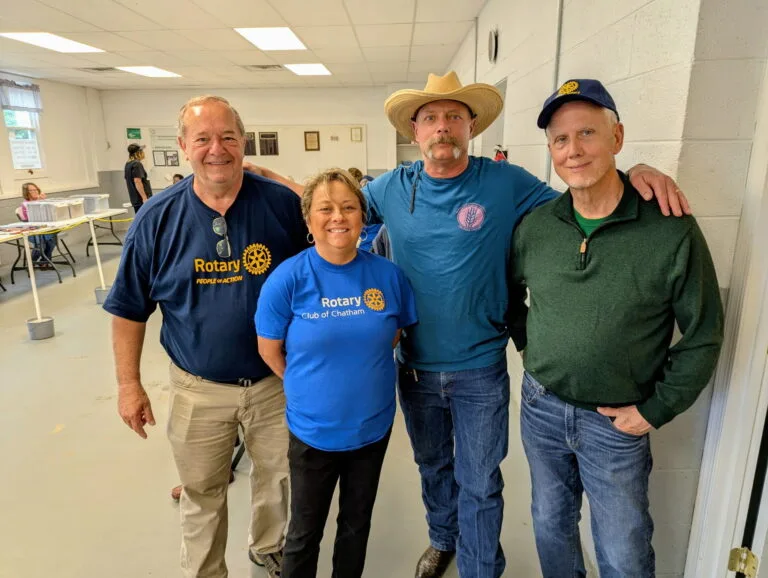 May show: Four adults stand together indoors, two wearing Rotary Club shirts, one in a cowboy hat, and one in a green sweater, posing for a group photo.