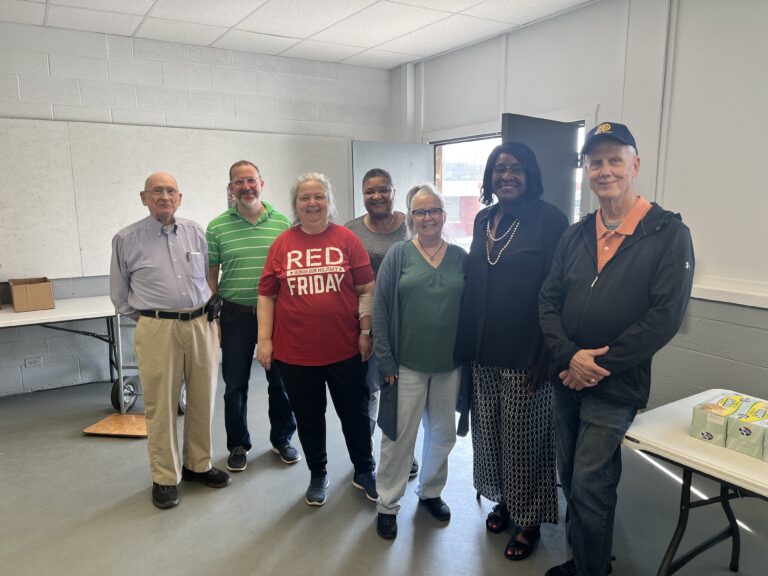 May show: Seven adults stand together in a room with tables, smiling at the camera. Some are casually dressed, and one wears a "Red Friday" shirt. The room appears to be a community space.