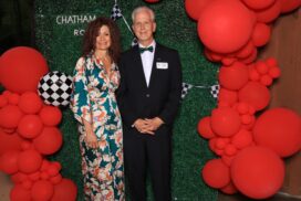 May show: A woman in a floral dress and a man in a black tuxedo stand in front of a green backdrop decorated with red balloons and checkered flags.