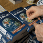 May show: A person places circular stickers on stacked copies of "A Student's Dictionary" on a wooden table.