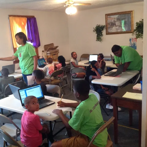 May show: A group of children work on laptops at tables in a classroom while two adults in green shirts assist them.