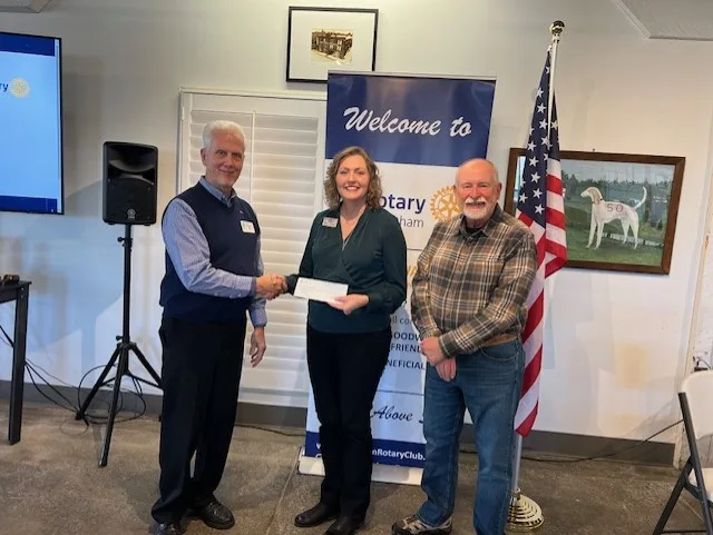 May show: Three people stand indoors; one man hands a check to a woman. They pose in front of a Rotary Club sign and an American flag. Another man stands to the right, smiling.