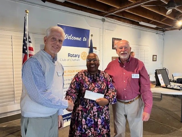 May show: Three people stand indoors, smiling, as one hands a check to another. A Rotary Club of Chatham banner is visible in the background.