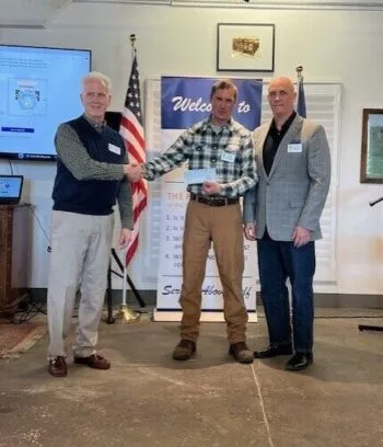 May show: Three men pose indoors; two shake hands while one stands beside them. An American flag and "Welcome to" banner are visible in the background.