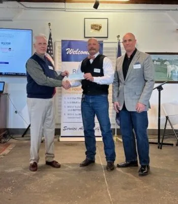 May show: Three men stand indoors; two of them shake hands while holding a check. A banner, flags, and a television screen are visible in the background.