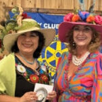 May show: Two women in colorful dresses and decorative hats stand smiling indoors at an event; musicians and flags are visible in the background.