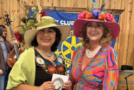 May show: Two women in colorful dresses and decorative hats stand smiling indoors at an event; musicians and flags are visible in the background.