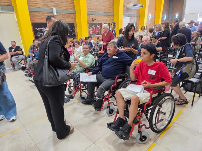 May show: A group of people, including individuals in wheelchairs, gather indoors for an event; some are seated, some are standing and talking, while one person eats a snack.