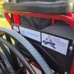 May show: Close-up of a red wheelchair showing foundation and Rotary Club stickers on the armrest, with grass visible in the background.