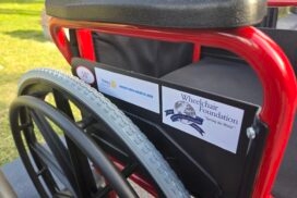 May show: Close-up of a red wheelchair showing foundation and Rotary Club stickers on the armrest, with grass visible in the background.