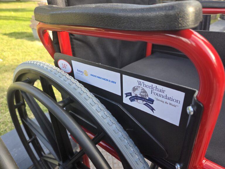 May show: Close-up of a red wheelchair showing foundation and Rotary Club stickers on the armrest, with grass visible in the background.