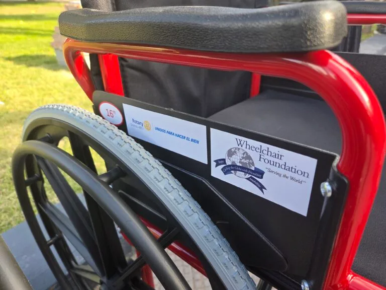 May show: Close-up of a red wheelchair showing foundation and Rotary Club stickers on the armrest, with grass visible in the background.